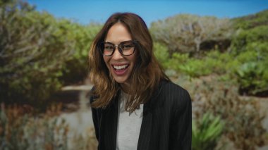 Parkta gülen bir kadın yemyeşil gözlüklü ve spor ceketli neşeli ve enerjik bir atmosfer taşıyor..