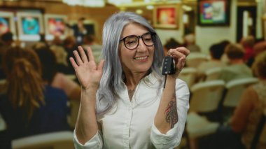 Sanat galerisinde elinde anahtarlarla gülümseyen gri saçlı yaşlı bir kadın. Etrafı insanlarla ve renkli resimlerle çevrili..