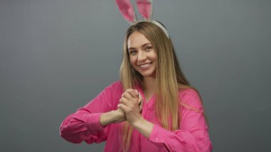 Uzun saçlı ve paskalya tavşanı kulaklı bir kadın izole edilmiş gri bir arka plan karşısında neşeli bir şekilde gülümsüyor, canlı pembe bir gömlek giyiyor, neşeli bir tatil ruhunu somutlaştırıyor..
