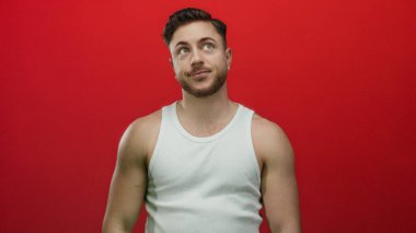 Sakallı ve kolsuz tişörtlü genç adam canlı kırmızı arka planda düşünceli ve rahat görünüyor. Gözleri kapalı, sıradan moda ve kendine güveni var.