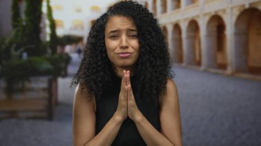 Eski bir kasaba sokağında kıvırcık saçlı bir kadın güneşli bir ortamda umut dolu bir ifadeyle ellerini sıkar..