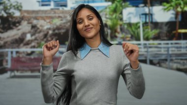 İspanyol kadın canlı bir sokakta arka planda tropikal manzarayla dans ediyor şık bir süveter giyiyor ve büyüleyici bir gülümseme sergiliyor..