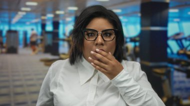 Gözlüklü kadın şaşırmış bir ifadeyle spor salonunda dikiliyor. Etrafı bulanık spor aletleri ve spor salonu geçmişiyle çevrili. Şok geçiriyor ya da inançsızlaşıyor..