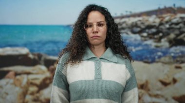 Gözlüklü bir kadın berrak bir gökyüzünün altında parlak mavi bir denizle kayalık bir sahilde duruyor, dingin ama düşünceli bir hava yayıyor..
