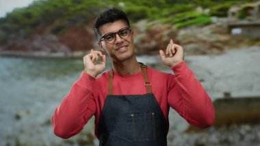 Gözlüklü ve önlüklü genç adam sahilde neşeyle gülümsüyor manzaralı bir arka planda.