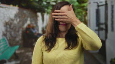 Kadın açık havada canlı bir şehir ortamında, neşeli ve samimi bir şekilde yüzünü kapatıyor. Sahne arkası banklar, yeşillik ve dokuma duvarlar var..