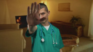 Steteskoplu yeşil üniformalı bir doktor bir otel odasında durup el hareketi yapıyor. Gayri resmi bir ortamda ihtiyat duygusunu ya da inkâr duygusunu vurguluyor..