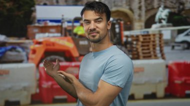 Mavi gömlekli genç adam inşaat alanında jest yapıyor. Arka planda ekipman ve bariyerler var. Gün boyunca..