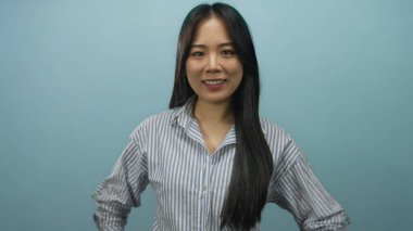 Mavi bir duvara karşı güvenle poz veren, çizgili bir gömlek giyen, neşeli ve rahat bir ruh hali taşıyan, elleri kalçasında ve göz teması kuran bir kadın..