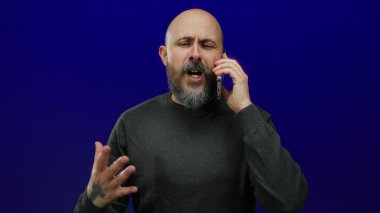 Kel ve sakallı bir adam cep telefonuyla izole edilmiş mavi bir arkaplan karşısında konuşuyor koyu renk bir gömlek giyiyor ve el hareketleriyle kaygılarını açıkça ifade ediyor.