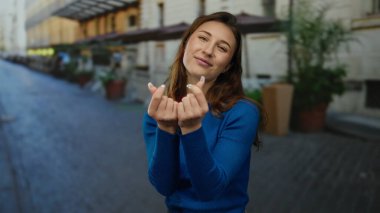 Genç bir kadın şehir sokaklarında gülümserken parmaklarıyla kalp hareketi yapıyor, şehir ortamında mutluluğu ve bağlantıyı ifade ediyor..