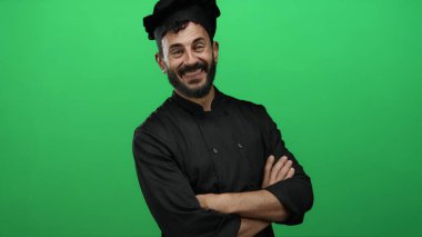 Siyah üniformalı, gülümseyen İspanyol erkek şef canlı yeşil arka planda aşçılık uzmanlığına karşı güvenle duruyor..