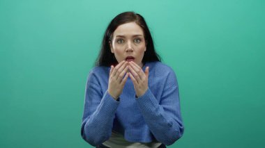 Şaşkınlık ve şaşkınlık ifade eden bir kadın canlı yeşil bir arka plana karşı, mavi bir kazak giyen, esmer oyuncu ve meraklı duyguları vurgulayan, geniş gözlü, eller kaldırılmış.