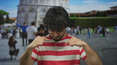 Güneşli bir günde sidik kulesinin önüne işaret eden gözlüklü ve sakallı İspanyol genç adam İtalya 'da turist ve mimariyle çevrili..