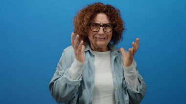 Mavi bir arkaplan önünde korku ve endişeyi ifade eden orta yaşlı bir kadın, duygusal ifadelere ve jestlere odaklanıyor, stüdyo ortamında gerginlik ve stresi canlandırıyor..