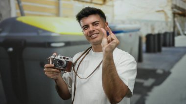 Şehir sokağındaki İspanyol asıllı genç adam elinde antika bir kamera tutarken parmaklarıyla şakacı jestler yapıyor ve canlı şehir sahnesini yakalıyor..