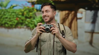 Elinde kamera ve sırt çantası olan beyaz sakallı genç bir adam, canlı bir şehir caddesinde fotoğraf yolculuğunun tadını çıkarıyor..