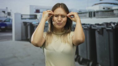 Kadın şaşkınlığını gözler kapalı ve elleri şehir sokağında yüzlerine yakın ve arka planda çöp kutularıyla ifade ediyor..