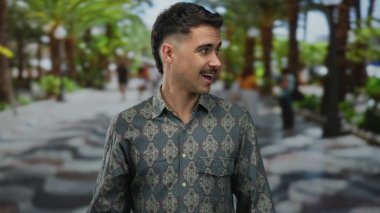 Şehir merkezindeki Latin kökenli genç adam, şehir merkezinde bulanık yayalar ve palmiye ağaçlarıyla çevrili, kendine güvenen bir ifadeyle desenli gömlek giyiyor..