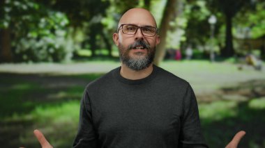 Gözlüklü, sakallı ve dövmeli bir adam güneşli bir park ortamında gülümsüyor, yemyeşil ortamda rahat ve kendinden emin bir tavır sergiliyor..