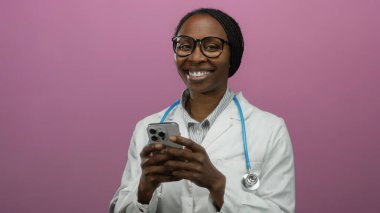 Doktor gülümserken pembe arka planda cep telefonu kullanıyor, steteskop takıyor, büyük gözlük takıyor, beyaz önlük takıyor, profesyonel iletişim ve mutluluk taşıyor.