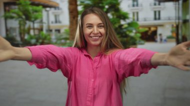 Pembe tişörtlü, kendini kucaklayan, bulanık geçmişiyle şehir merkezinde neşeli bir çevre ve canlı bir yeşillik öneren bir kadın..