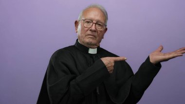 Siyah cüppeli kıdemli rahip, mor arka plana karşı eli açık el kol hareketi yapıyor..