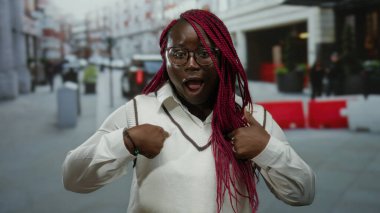 Afro-Amerikalı bir kadın, hareketli bir şehir caddesinde hareketli bir şehir manzarası sergileyerek kendine sürpriz yüz ifadeleri sergiliyor..