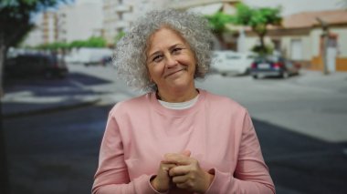 Güneşli bir günde, şehir caddesinde gri kıvırcık saçlı, pembe kazaklı, sıcacık gülümseyen bir kadın..