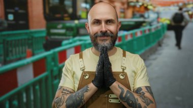 Dövmeli ve sakallı kel adam inşaat alanında gözleri kapalı eldiven ve tulum giyiyor..