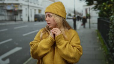 Sarı kazaklı ve şapkalı kadın şehir ortamında korkuyu yakalayan açık hava ortamını tasvir eden şehir ortamında endişeli görünüyor..