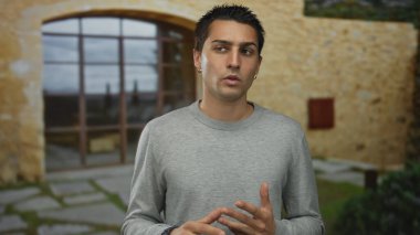 Geniş pencereli, taşra tuğlalı bir binanın yanında gündelik kıyafetlerle dikilen genç İspanyol adam düşünceli ve sohbete dalmış görünüyor..