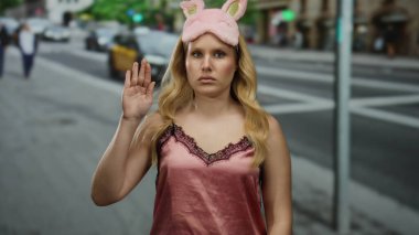 Pembe gecelikli ve uyku maskeli genç sarışın kadın gündüz vakti şehir sokaklarında dikiliyor..