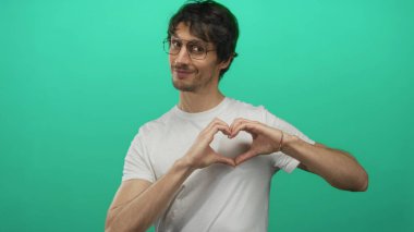 Gözlüklü ve beyaz tişörtlü genç adam yeşil stüdyoda elleri ile bir kalp şekli yapıyor..