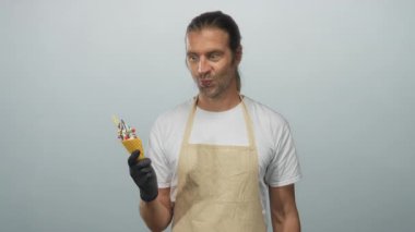 Önlük takan uzun saçlı sarışın adam açık gri stüdyo ortamında siyah eldivenli dondurma külahını tutuyor..