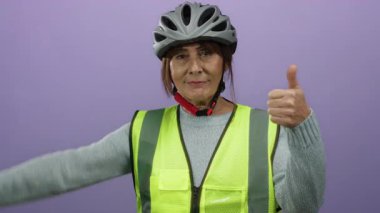 Güvenlik yeleği ve miğfer giyen kıdemli İspanyol kadın mor bir duvara karşı duruyor, iki başparmağını da öne sürüyor, karışık duygular ya da zıt görüşler öne sürüyor..