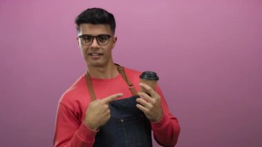 Kırmızı süveter ve önlük giymiş, pembe arka planda kahve tutan ve bardaklarla gülümseyen bir adam neşeli ve davetkar bir jest yapıyor..