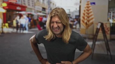 Uzun saçlı sarışın adam caddede karnını tutarken yüzünü ekşitiyor; acı mide bulantısı sıkıntısı.