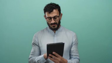 Sakallı ve gözlüklü İspanyol bir adam yeşil arka planda tablet kullanıyor, mavi gömlek giyiyor ve odaklanmış ve düşünceli görünüyor..