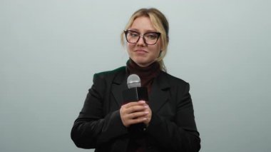 Mikrofonlu bir kadın beyaz bir duvara yaslanmış gözlüklü ve takım elbiseli bir kadın belirsizlik ya da soru işareti taşıyarak ifade verirken rapor veriyor..