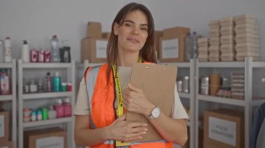 Yansıtıcı yelek giyen bir kadın bağış merkezinde pano tutuyor. Arkaplanda raflar var. Kapalı alanda gönüllü bir çalışma alanı var..