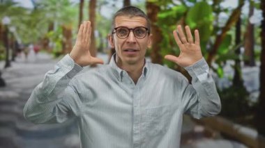 Palmiye ağaçları ile çevrili, şaşırmış ifade ve gözlüklü adam, bir şok ve merak anı yakalar gelişigüzel şehir ortamında.