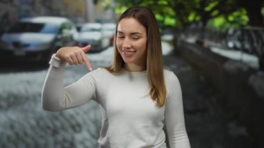 Sokakta beyaz kazaklı genç bir kadın park etmiş arabalarla ve şehir merkezinde yeşil ağaçlarla çevrili neşeli bir ifadeyle..