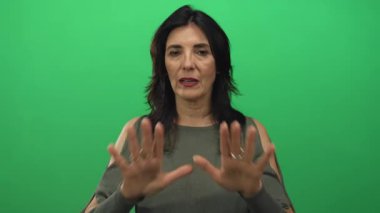 Yeşil bluzlu İspanyol kadın, stüdyo ortamında ciddi bir ifadeyle avuçlarını ileri açıyor..