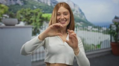 Okyanus tepelerine tepeden bakan balkonda elleri olan kalp şeklinde genç sarışın bir kadın..