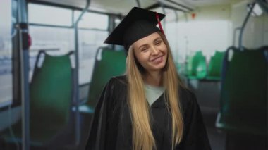 Mezuniyet elbisesi giymiş, gülümseyen ve otobüste öpüşen genç bir kadın beklenmedik bir yerde kutlama ve başarı simgeliyor..