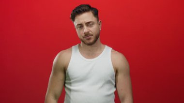 Sakallı ve kolsuz tişörtlü genç adam canlı kırmızı arka planda düşünceli ve rahat görünüyor. Gözleri kapalı, sıradan moda ve kendine güveni var.