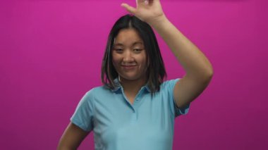 Çinli kadın canlı pembe arka planda ezik jestler yapıyor. Mavi gömlekli, neşeli ifadeli ve kendine güvenen bir duruş takınıyor..