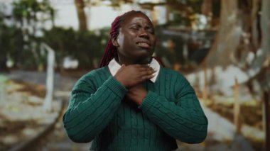 Yeşil kazaklı bir kadın parkta gergin bir ifadeyle boynuna dokunuyor. Bulanık ağaçlar ve patikalarla çevrili nefes almakta güçlük çekiyor..