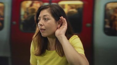 Bir yeraltı binasının içinde, bir metro treninin yanında eli kulağında oturan bir kadın anonsu duymak için öne eğiliyor:.